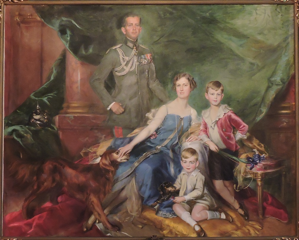 Op de achtergrond een interieur met marmeren zuilen met daarvoor een groen gordijn. Net links van het midden van het portret staat een man in een groen militair uniform. In het midden schuin zittend een dame met kort haar, oorbellen in en een sierlijke blauwe avondjurk. Rechts naast haar staat een jongen met een witte blouse met een fladderige kraag, een roze-rood jasje en een zwarte kortebroek. Hij heeft zijn linkerhand in zijn zij, met zijn rechter arm leunt hij op dezelfde leuning als zijn moeder. In zijn rechterhand heeft hij een iris. Voor op de grond zit een kleinere jongen met een witte blouse, een beige jack en korte broek. Hij houdt een teckel vast die naast hem, op de jurk van zijn moeder, ligt. De dame heeft haar rechter hand op de snuit van een Ierse Setter. Helemaal links ligt op het groene gordijn een pickelhaube, een duitse militaire helm met een punt erop.
