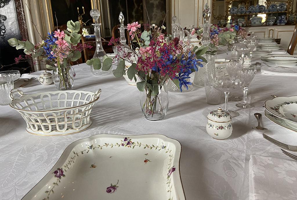 Een feestelijk gedekte tafel met veel bloemen. Op de voorgrond een rechthoekige schaal met een bloemenrand en losse bloemen. Links daarnaast een mandje van porselein, rechts een dekselpotje.