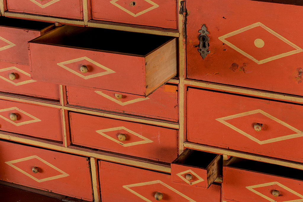 Een kast met allemaal verschillende formaten lades, een paar lades staan half open. De lades hebben een rode voorkant met een klein geel rond knopje waar omheen een liggende gele wieber is geschilderd. De lade rechts boven is waarschijnlijk een deurtje, want dat heeft een slot.