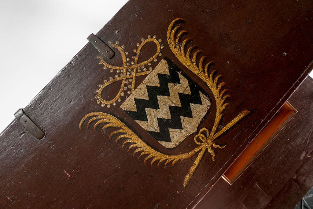 Bovenkant van de reiskist van Godard Adriaan van Reede. De bovenkant is schuin gefotografeerd, de kist is donkerbruin, aan de bovenkant zitten twee meegeverfde sluitingen. Op de kist is het familiewapen van de Van Reedes geschilderd: wit met een zwart witte zigzag. Om het wapen heen zitten twee gouden palmbladeren en er boven een sierlijke krul met witte stippen (parels?). Rechts onder zie je nog een lade open staan.