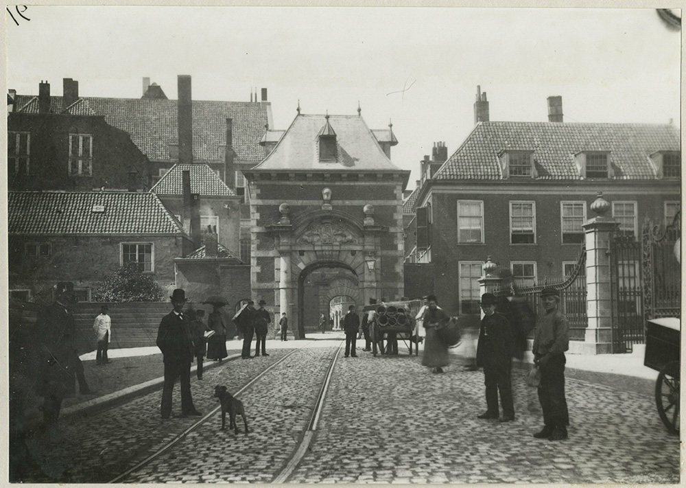 Straat richting de Mauritspoort van het binnenhof. De straat is geplaveid met kinderkopjes en er lopen tramrails. Diverse mensen lopen op straat heren met bolhoed, jongens met pet en dames. In het gebouw naast de mauritspoort staan de ramen open. de gebouwen achter de mauritspoort hebben hoge schoorstenen.