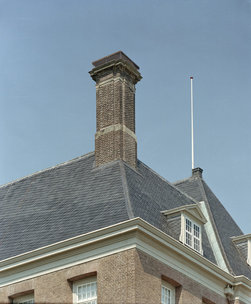 Van onderaf zoen we schoorsteen op de hoek van een leien dak. Onder de schoorsteen een raam en achter de schoorsteen op de nog van het hoogste dak een vlaggenmast.