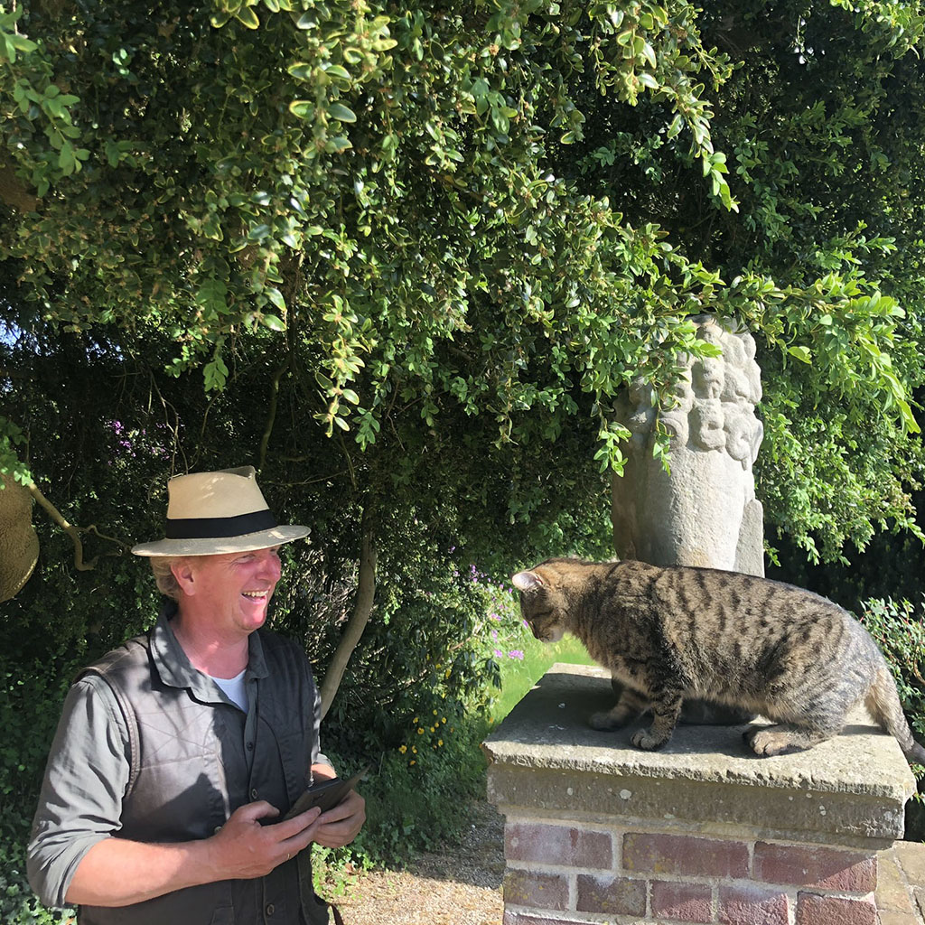 Onder een boom staat een bakstenen pilaar met een hardstenen klapmuts. Op de klapmuts staat een stenen leeuw met zijn rug naar ons toe, daarvoor zit een cyperse kat. Naast de pilaar staat een man met een hoed op die lacht naar de kat.