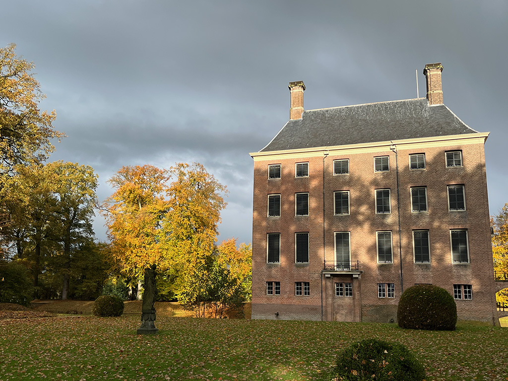 Kasteel Amerongen tegen een heel erg donkere lucht. De zon schijnt, op het huis zijnd de schaduwen van bomen te zien. Op het grasveld op de voorgrond liggen blaadjes, de blaadjes aan de bomen zijn geel tot roestbruin.