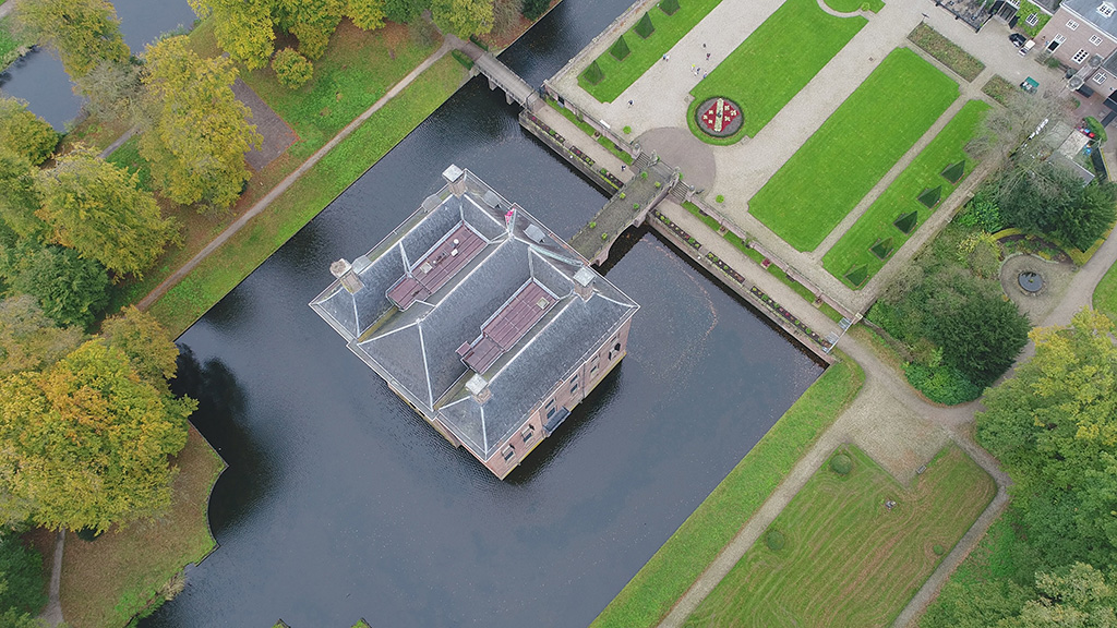 Foto van bijna recht boven kasteel Amerongen. In een vierkante gracht ligt een vierkant huis. Er gaat één brug naar het huis toe. Het huis heeft drie daken en voor zijn ze verbonden tot één dak. Aan de kant van de brug is een plein met groene grasvelden, een bloemenperk met het wapen van de heerlijkheid Amerongen. Aan weerszijden van het plein staan scherp geschoren taxuspyramides als pionnen op een schaakbord. Linksboven en rechts bomen, verder vooral gras en paden.