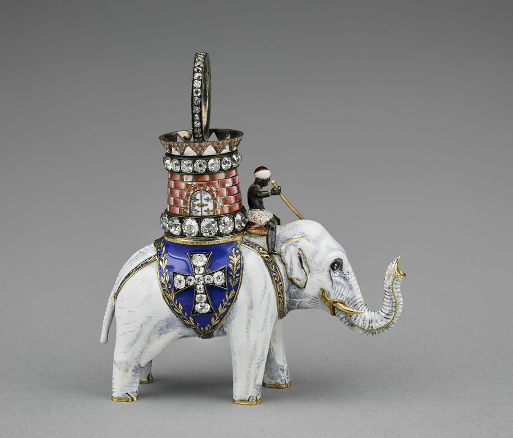 Een hanger van een witte olifant met goud afgezet. Achter de oren zit een zwarte man die de olifant bestuurd. De olifant draagt een blauw dekje met daarop een kruis van diamanten. Op zijn rus heeft hij een rode ronde toren ook bezet met diamanten.