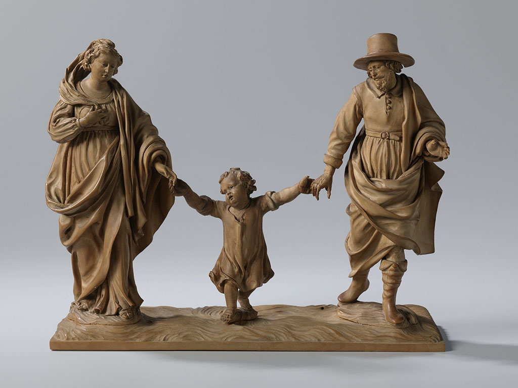 Deze beeldengroep van een vader, moeder en kind, stelt de Heilige familie voor. Op een naturalistische ondergrond wandelen Maria en Jozef zij aan zij met hun zoon tussen hen in. Het paar houdt de handen van het Christuskind vast om hem te helpen bij het lopen. Ze lijken op het punt te staan de peuter speels een stukje omhoog te zwiepen. Maria draagt onder een omslagdoek een gewaad met ceintuur hoog rond het middel. Ze loopt op sandalen en in haar gevlochten, opgestoken kapsel is een hoofddoek verwerkt. Jozef draagt eigentijdse kleding, laarzen en een hoed met een riem. Om zijn rechterschouder heeft hij een mantel geslagen. Het Christuskind loopt op blote voeten en draagt een eigentijds hemd met opgestroopte mouwen. De ondergrond en de drie figuren zijn separaat gesneden, daarnaast zijn de armen en Jozefs hoed los aangezet. De ogen van de drie figuren zijn ingelegd met zwart glas.