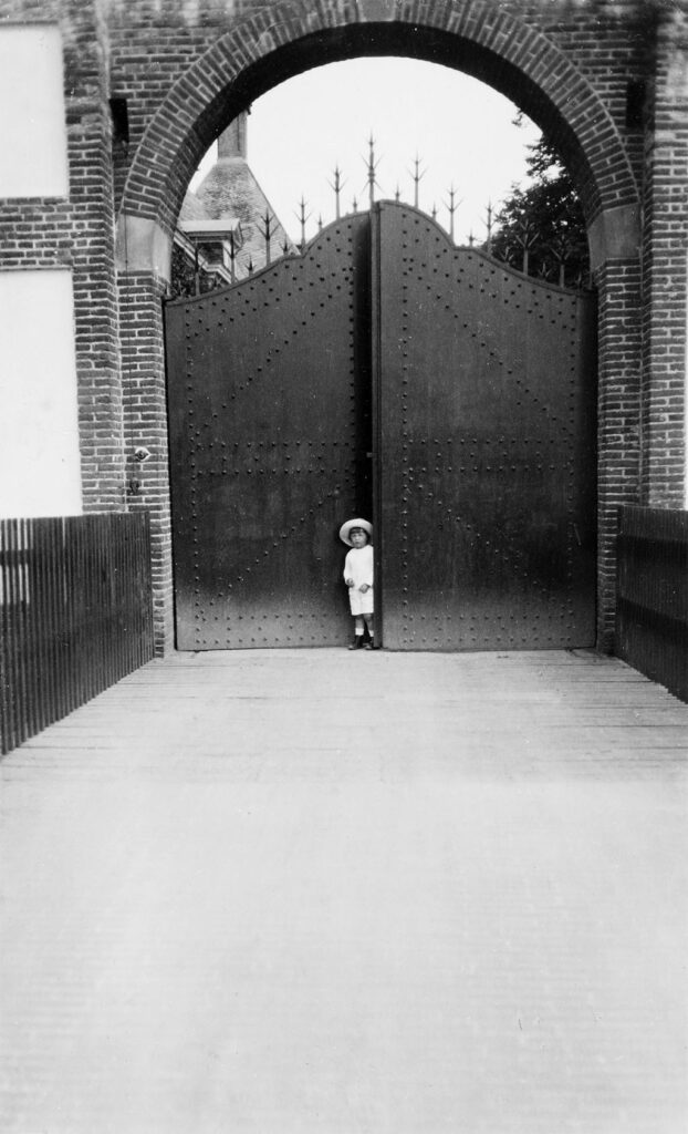 Tussen de twee deuren van een grote poort staat een kleuter met een wit shirt en een witte korte broek, witte sokken en zwarte schoenen. Hij heeft een witte hoed op. De deuren zijn hoog en door flinke spijkers is de versteviging aan de andere kant goed te zien. Op de deuren staan grote stekels.