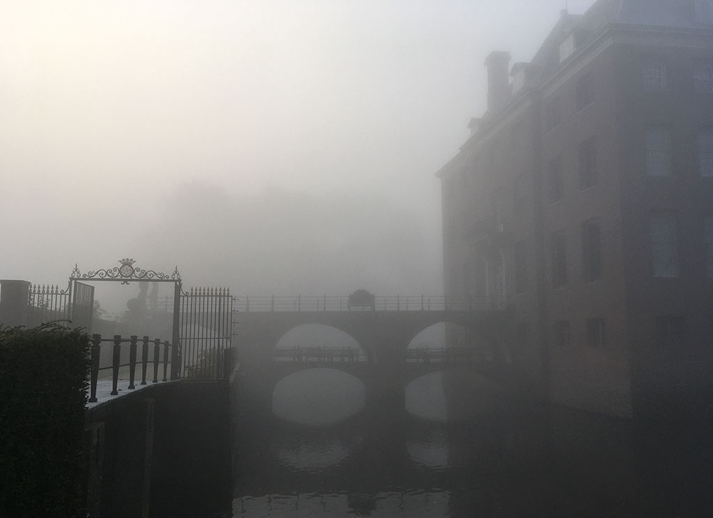 Een mistige foto van het kasteel. Rechts het kasteel dat nog vaag zichtbaar is. De dubbele brug is goed te onderscheiden en ook de weerspiegeling daarvan in de gracht. Links een brug en een hek, op de achtergrond zijn nog heel vaag de contouren van bomen te zien.