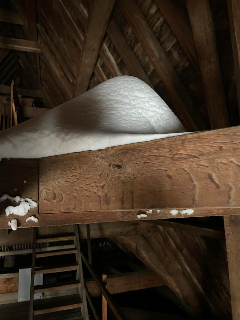 Op de balken van de zolder ligt een bergje sneeuw. Op de achtergrond de dakspanten en een houten trap