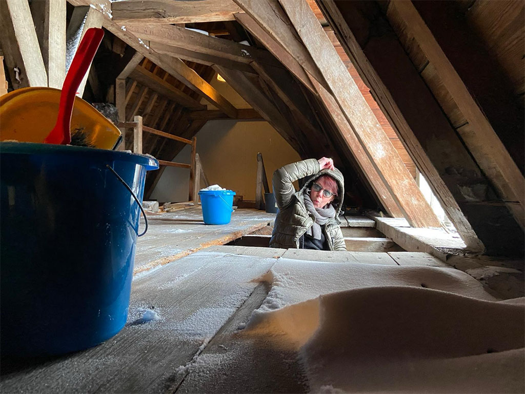 Op de zolder ligt sneeu op een houten vloer. Voor het raam zit iemand met een muts op. Her en der staan emmertjes met vegers en sneeuw.