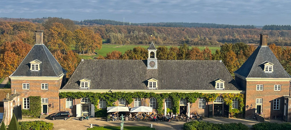 in het midden een breed gebouw met hoge boogramen en -deuren. Het heeft geen verdieping maar in het pannendak zitten drie koeken. In het midden staat een klokkenstoel. Aan weerszijde van het lange gebouw staat een vierkant gebouw met twee verdiepingen en een pyramidedak met leien. Elk dak heeft een koekoek en boven op de punt staat en schoorsteen. Voor het middelste gebouw ligt een vol terras waarom de mensen genieten van de laagstaande avondzon. Op de achtergrond bomen in herfstkleuren rondom een weidelandschap. Helemaal op de achtergrond de glooiing van de Utrechtse Heuvelrug.