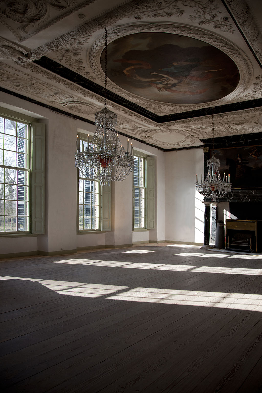Foto van een helemaal lege grote zaal. Er staan geen meubels, maar er hangen wel kroonluchters. De luiken zijn open en de zon schijnt op de houten vloer.