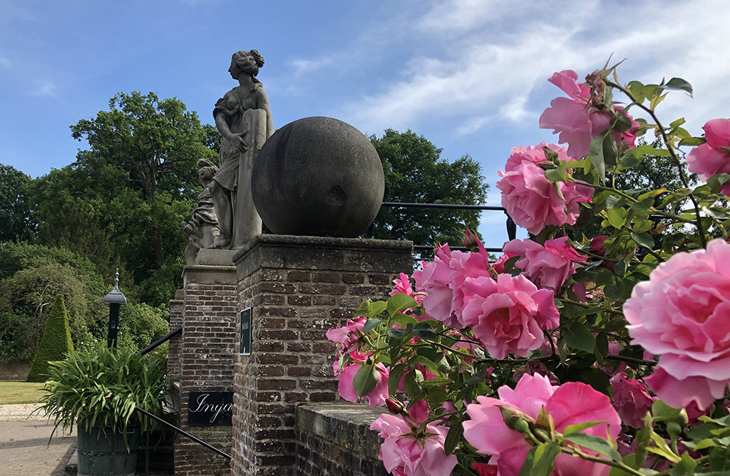 Foto van het bordes van Kasteel Amerongen vanaf de zijkant. Op de voorgrond rozen die over de muur groeien, daarachter een bal op een pilaar. Achter de bal een standbeeld van een vrouw met een pilaar in haar hand. Achter haar nog een onduidelijk standbeeld.