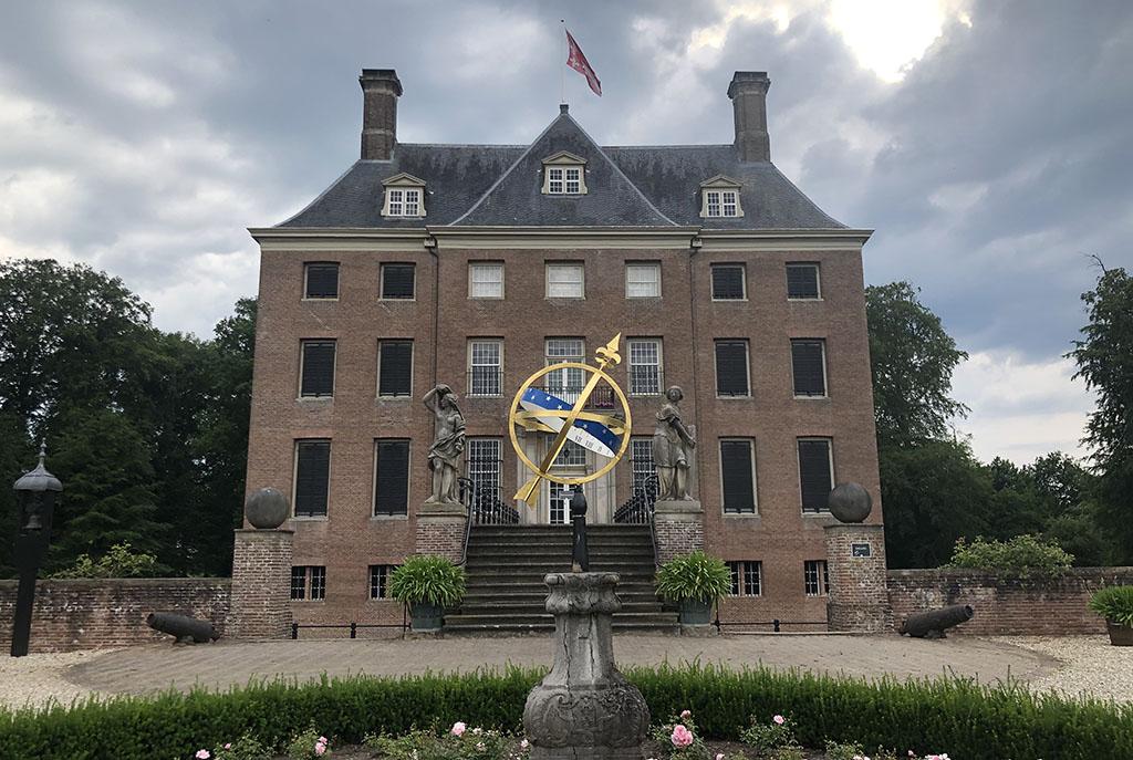Foto van Kasteel Amerongen. Een hoog, bakstenen vierkanten huis met twee schoorstenen. Voor het huis een trap naar een bordes. Aan weerszijden van de trap twee standbeelden. Daarnaast twee ruimtes en dan twee pilaren met daarop ronde ballen. De pilaren gaan over in een muur, waarover struiken groeien. Voor de pilaren twee kanonnen en helemaal op de voorgrond een gouden zonnenwijzer.