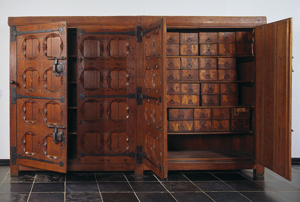 Archiefkast van eikenhout. Twee doorgaande hoekstijlen en een middenstijl dragen aan ijzeren kruishengsels vier opgeklampte deuren. De meest linker deur staat op een kier, de twee rechterdeuren staan wijd open, waardoor je de lades ziet. De door de kruising der klampen verkregen vierkanten zijn binnen ingekerfde cirkels Gotisch gebiljoend. Ieder paar deuren heeft twee ijzeren sloten met grendels. Een tap met een gat is aan de ene hoekstijl bevestigd en een draaibare beugel aan de andere. Binnenin veertig van de oorspronkelijke laden met Gotische majuskels.