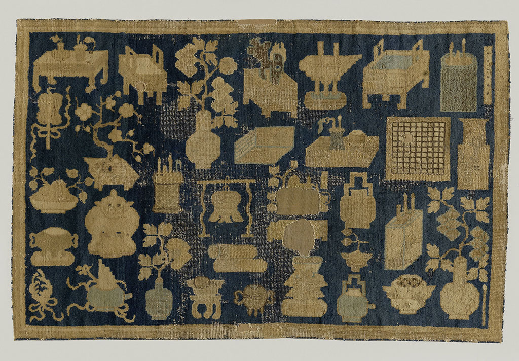 Oosters tapijt met de honderd antiquiteiten. Middenveld: een blauwe achtergrond is bedekt met rijen van de zogenaamde honderd antiquiteiten in beige, grijsblauw en bruin, waaronder een tafel, een bloesemtak in vierkante pot, een penseelpot, een waaier, een dambord, een bel, twee samengestrikte veren, een bolvormige vaas en een staande penseel. Rand is effen beige.