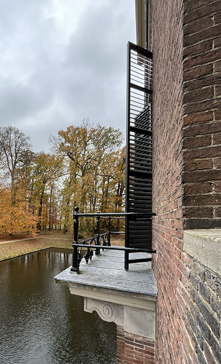 Foto langs de muur naar een balkon genomen. De luiken staan open. Het balkon is grijs met een zwart geschilderde metalen ballustrade.