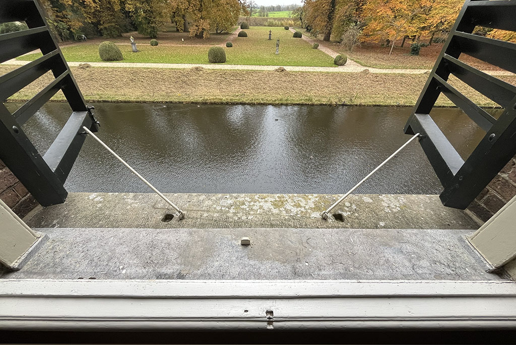 Foto van een open raam met twee open luiken. De haken waarmee de luiken openstaan zitten in een hardstenen vensterbank. Vanuit het raam zie je de gracht met een paar eendjes en op de achtergrond een grasveld met bolbuxussen en er omheen bomen in herfskleuren. 