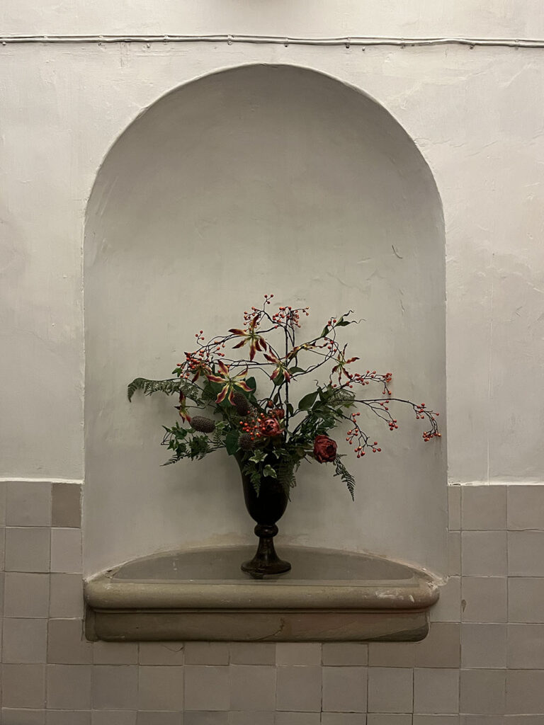 Een wit gestucte muur met een nis. Boven de nis lopen elektriciteitsdraden horizontaal, de muur aan de onderkant is betegeld met witjes. Onder in de nis een stenen bak. Op de stenen bak staat een vaas met bloemen.