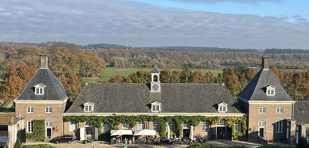 Foto van een breed gebouw van één verdieping met zadeldak met aan weerszijden twee paviljoens van twee verdiepingen met puntdak. Voor het gebouw is een terras waar de parasols open staan en mensen in het zonnetje zitten. Op de achtergrond de Utrechtse Heuvelrug in herfstkleuren. 