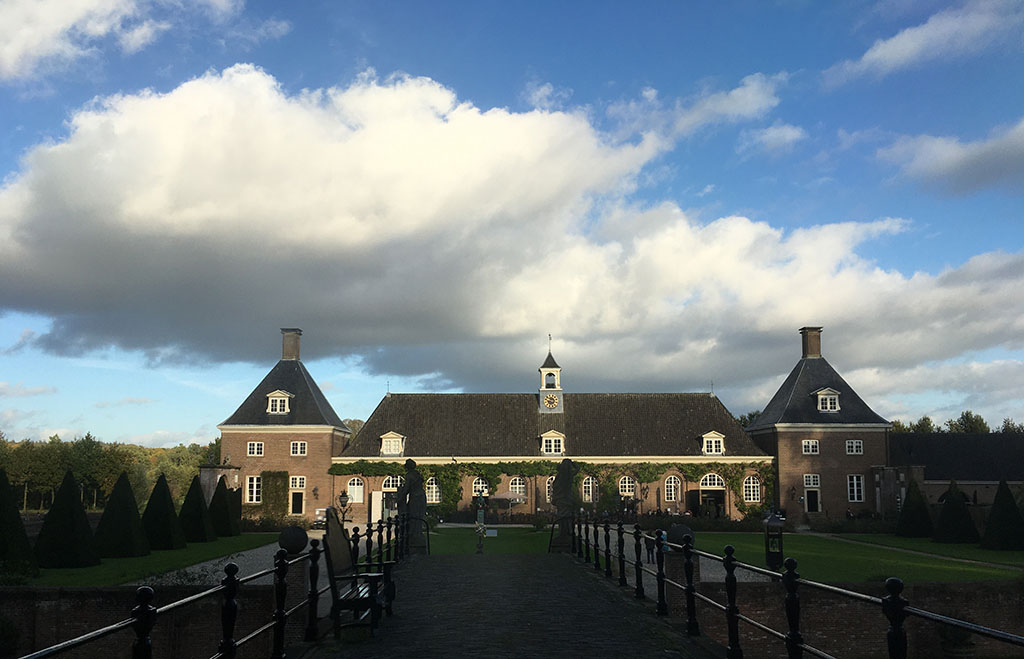 Foto vanaf de brug van het kasteel naar het oosten waar de stallen liggen. De stallen zijn een breed gebouw met een zadeldak en in het midden een klokkenstoel. Aan weerszijden staat een vierkant gebouw met een pyramidedak. Boven het stalgebouw hangt een grote wolk waarvan de onderkant grijs is en de bovenkant zwart. De zon schijnt op de stallen, de voorgrond is donker.