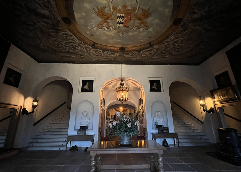 De hal van kasteel Amerongen. In het midden staat een tafel met daarop een boeket. Daarachter een gang met een hangende lamp in het midden van het gewelfde plafond. Aan weerszijde van de gang een trap naar boven. Tussen de trappen en de gang zit een nis met daarin een gipsen beeld en daarboven een nis met een schilderij. Naast beide trappen steekt een arm uit de muur die een lamp vast houdt. Het plafond is sierlijk beschilderd met in het midden het alliantiewapen van Godard Adriaan van Reede en Margaretha Turnor.