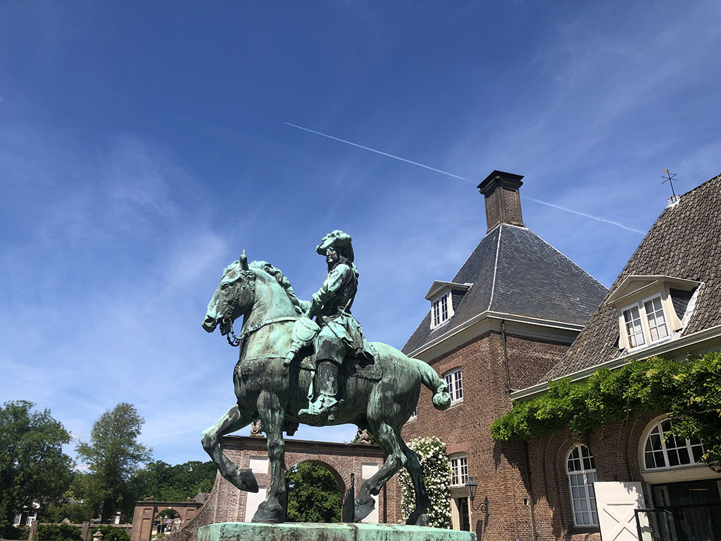 Foto van een ruiterstandbeeld van een man die zijn paard met ferme hand in toom houdt. De mond van het paard staat open. Het paard is in draf afgebeeld, de staart lijkt in beweging te zijn. De man is historisch gekleed. Hij draagt een hoed en laarzen met grote sporen.