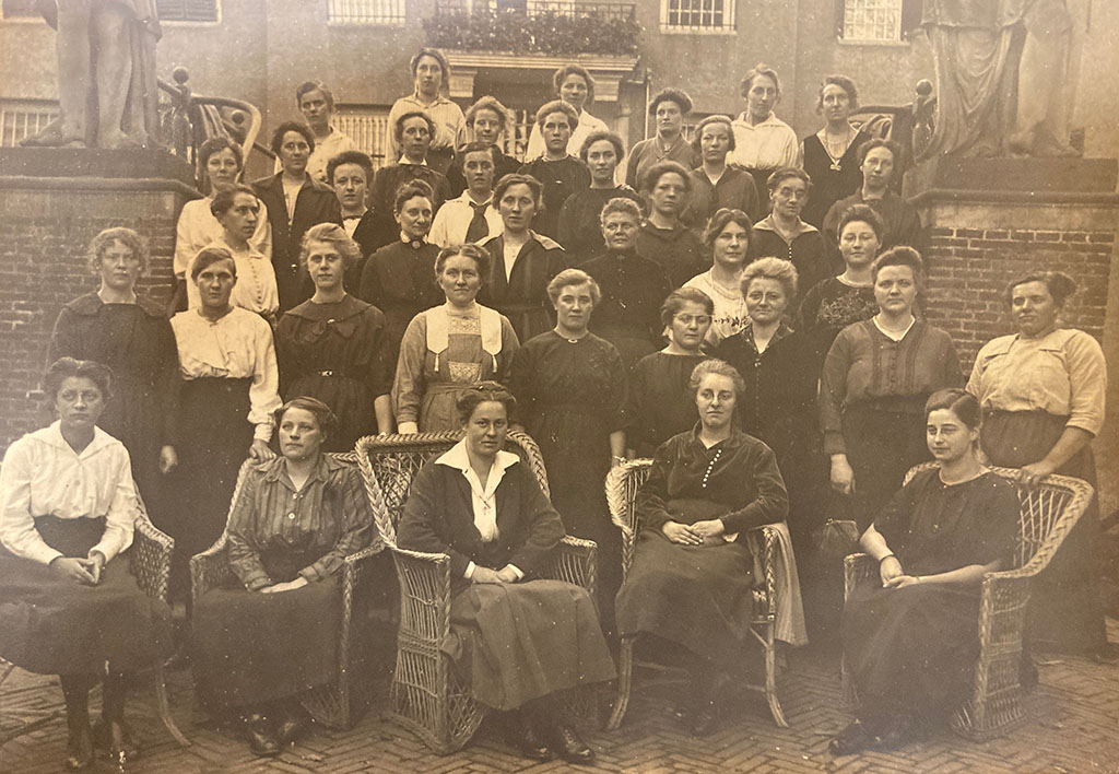Groepsfoto van netjes, maar eenvoudig geklede dames met opgestoken haren. voor de groep staan drie rieten stoelen waar ook dames in zitten.
