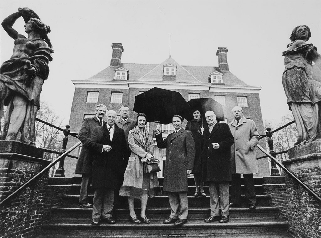 Tussen de standbeelden van Prudentia en Fortitude staan zes keurige heren en twee dames op de bordestrap. Ze hebben twee paraplu's bij zich.