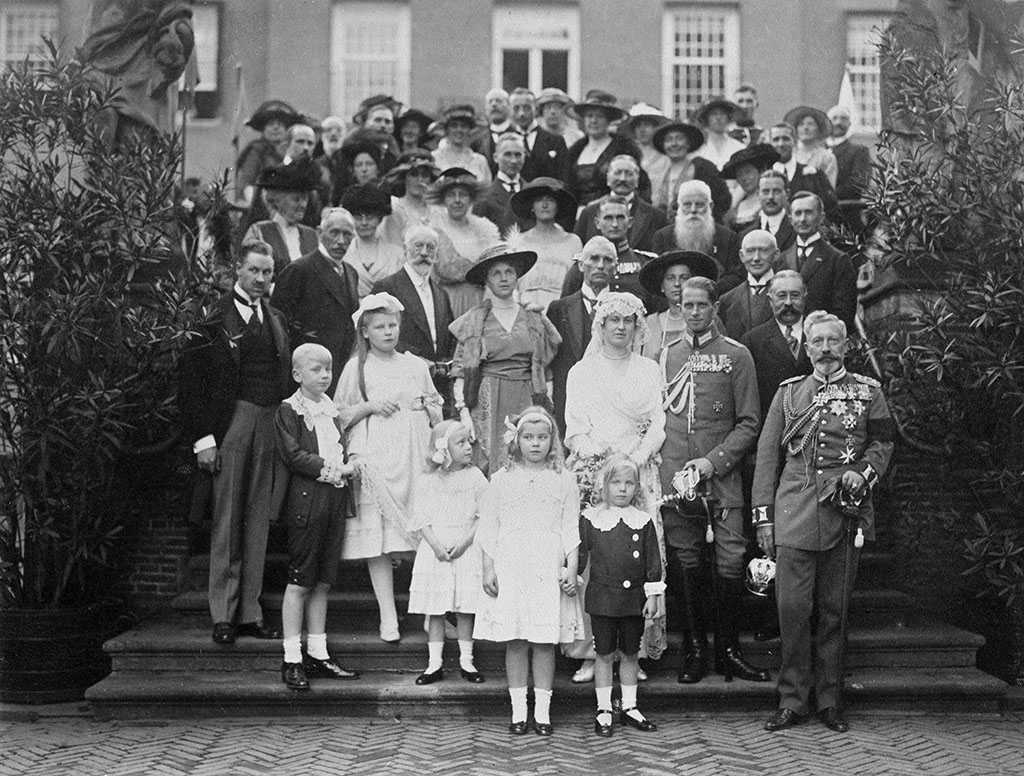 Bordesfoto van een huwelijk. In het midden de bruid in het wit met een kanten mutsje op. Naast haar de bruidegom in Duits uniform. Voor de foto verschillende kinderen, waarvan één meisje naar de bruid kijkt. Naast de bruidegom staat de vader van de bruid in pak, daarvoor de Duitse (ex-)keizer in uniform.