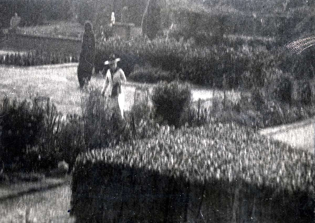 Een zeer bewogen foto van een tuin met borders en strak geschoren heggen. Op één van de paden loopt een dame in het wit in een getailleerde jas met een hoed op en een paraplu aan haar arm.