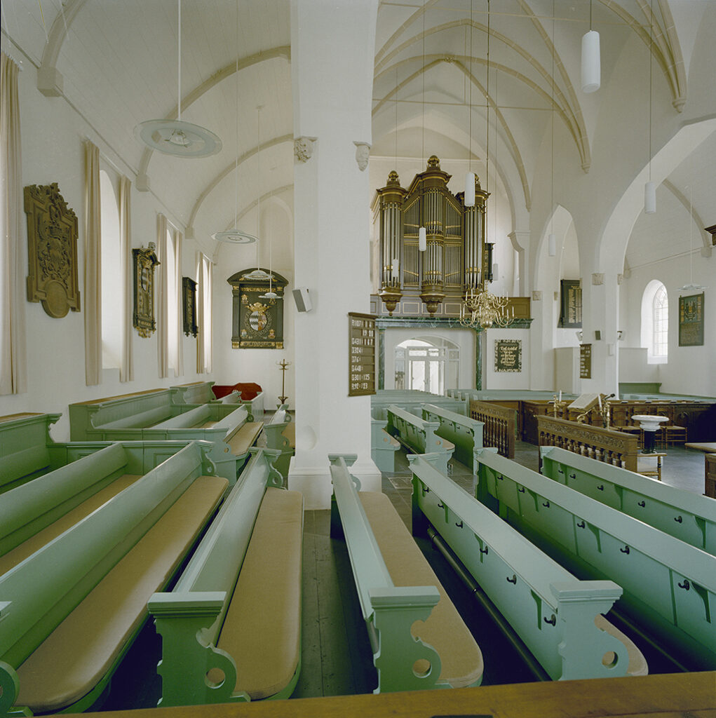 Interieur van een wit gestucte kerk naar de uitgang met daarboven het orgel. In de kerk staan groene banken en aan de muur hangen diverse rouwborden.