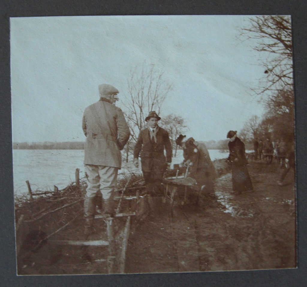 Op een modderig dijkje waar het water tegen de kant aan klotst zijn allemaal aan het werk. In de modder zijn palen in de grond geslagen en worden vakken gevuld met zand. In de modder staan een kruiwagen en dames in lange jurken.