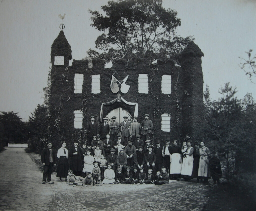 Op de splitsing van een weg is een groot kasteel nagebouwd met op de linker toren een haantje en een klok. Voor het bouwwerk staan dorpsbewoners te poseren. Op de voorgrond zitten kinderen, dan de vrouwen en een paar mannen en op een verhoging meer mannen. Het bouwwerk moet acht tot tien meter hoog zijn.