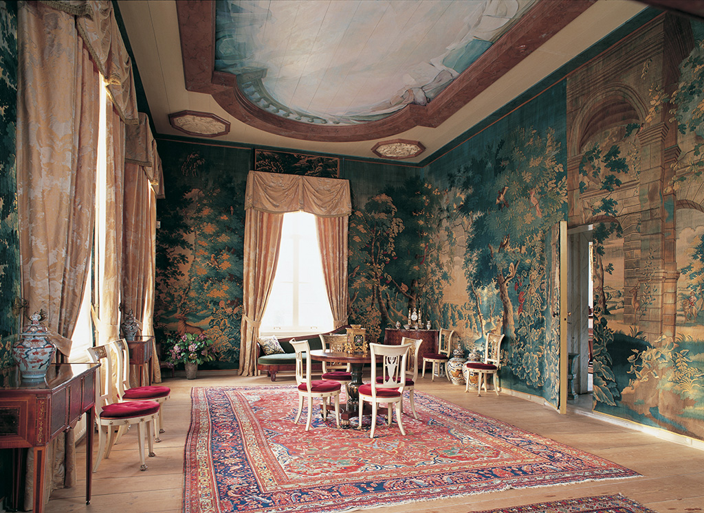 Een elegante kamer volledig behangen met wandtapijten. Rechts staat een (eveneens met tapijt beklede) deur op een kier. Midden in de kamer staat een ronde tafel met daaromheen vier stoelen. Voor het raam erachter staat een bank, in de hoek een comode. Links een wandtafeltje met daarop een Chinese fase. Langs de wanden staan nog vier witte stoelen. Op de houten vloer ligt een Perzisch tapijt. Het plafond is beschilder met een modern trompe l’oeuil