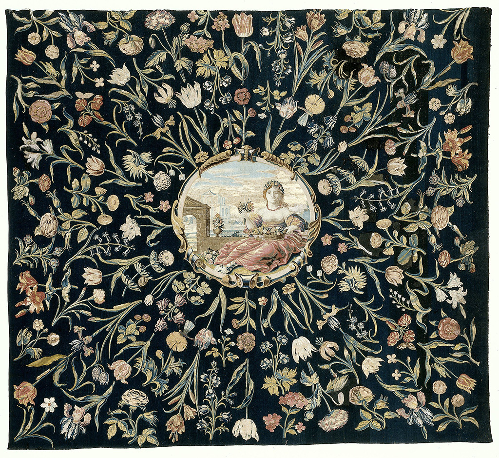 Tafelkleed van tapisserie-weefsel met in het midden Flora, Romeins godin van de lente en de bloemen, binnen een cartouche en daaromheen gestrooide bloemen op een donkerblauw fond.