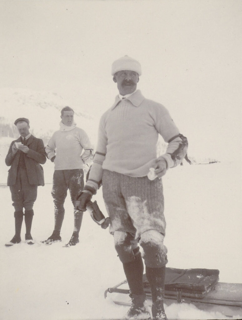 Een man met een snor staat in de sneeuw. Hij draagt een witte trui met armbeschermers, een kniebroek met lange kousen. In zijn hand heeft hij een zakdoekje.