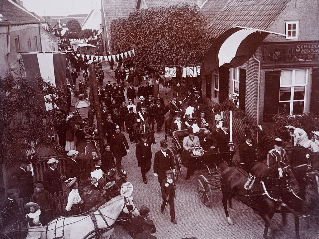 Zwartwit foto vanaf een hoog punt. We kijken een dorpsstraat in waar vlaggen uitgestoken zijn en vlaggetjes boven de weg hangen. Een koets voorgetrokken met twee paarden rijdt rechts onder het beeld uit. Op het paard links voor zit een heer een livrei, achter op de koets nog twee mensen in uniform. In de koets zitten een dame en een heer, de heer neemt net zijn hoge hoed af. Op straat zijn veel mensen die allemaal naar de koets kijken. Achter de koets rijden twee paarden, naast de koets lopen heren in pak. Erachter lopen allemaal mensen.