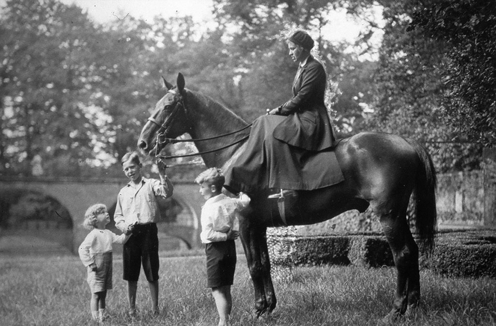 Op een groot paard zit een dame in dames zit met een rok en een jas aan. Het paard staan in een tuin: op de achtergrond zien we geschoren buxushaagjes. Bij het paard staan drie jongens. De oudste kijkt naar de fotograaf. Hij is een jonge teenager, houdt één van de teurgels vast met zijn linker hand, aan zijn rechterhand heeft hij zijn broertje. Hij is een jaar of vijf en heeft blonde krullen tot in zijn nek. Hij kijkt omhoog naar het paard en zijn moeder. De derde jongen staat naast het linker been van het paard. Zijn rechter hand ligt op de schouder van het paard, zijn linker hand heeft hij op zijn rug.