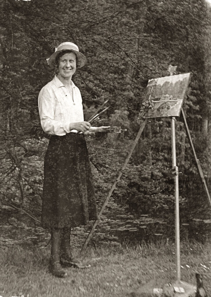 Zwartwit foto van een vrouw in een zwarte rok, een witte blouse en een zonnehoef op achter een schildersezel. In haar linker hand heeft ze een palet in haar rechter kwasten. Ze staat in het gras met op de achtergrond bomen. Op het schilderij zien we een brug.
