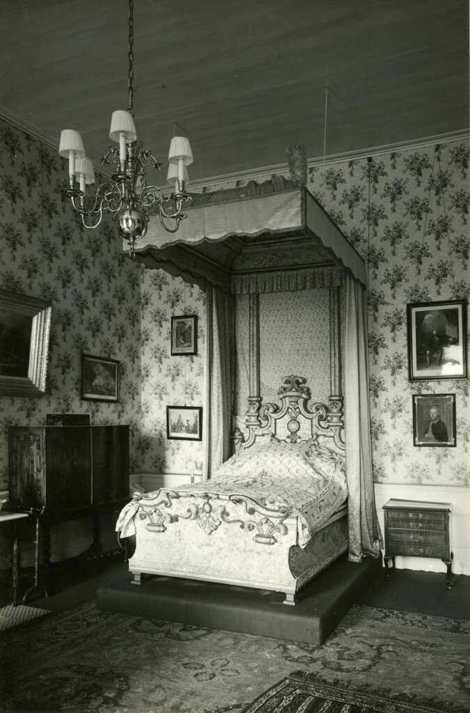 Zwarwit foto van een kamer met een behang met een regelmatig patroon. Op een kleine verhoging sataat een elegant versierd hemelbed.