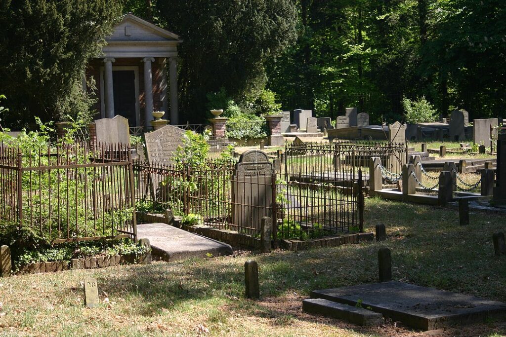Foto van een oude begraafplaats in het groen. Het gras is groen, maar op sommige graven staat onkruid. De diversiteit aan stenen en hekjes is groot. Op de achtergrond tussen de bomen staat een mausoleum met zuilen en een timpaan.