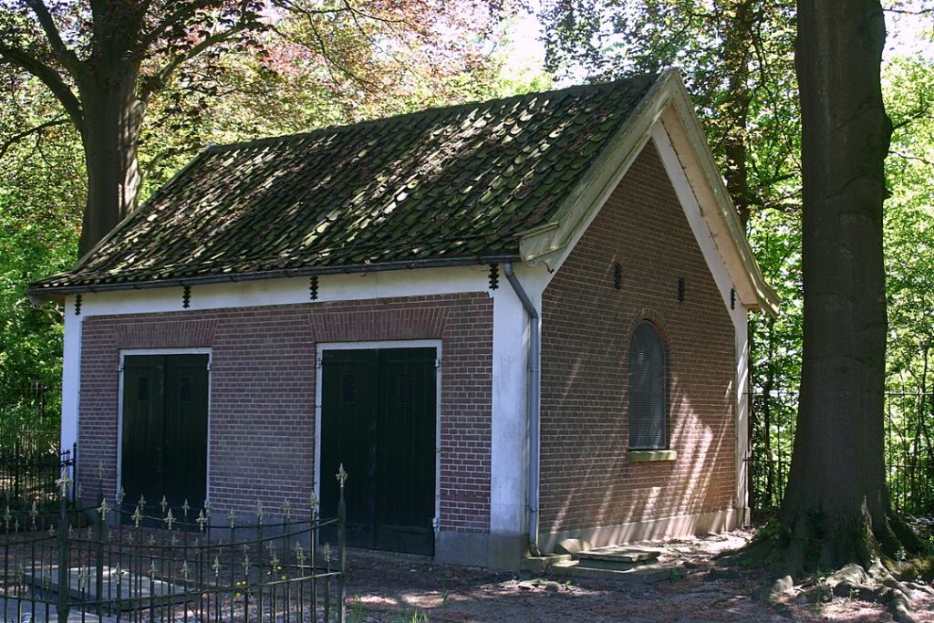 Een klein huisje met een puntdak in de schaduw van grote bomen. Aan de zijkant twee dubbele deuren, aan de kopse kant een raam met een boog.