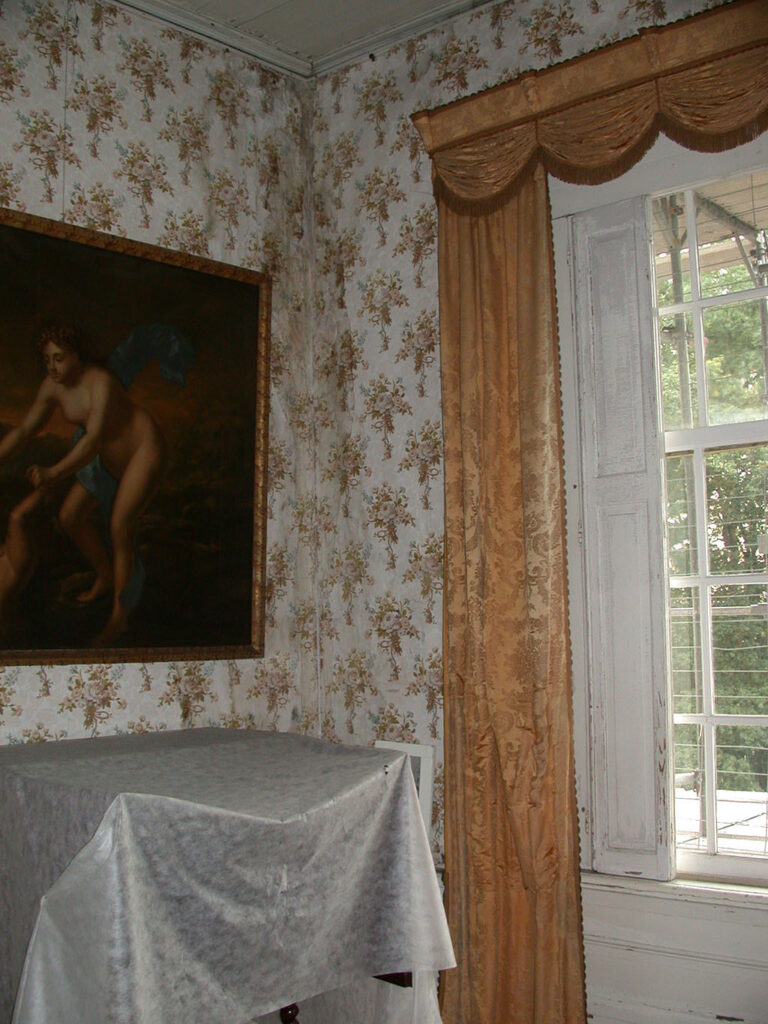 Hoek van een kamer met een bloemetjes behang. Rechts een raam met daarvoor een geel gordijn, links een schilderij met een naakte vrouw onder het schilderij een afgedekt meubelstuk. In de hoek is duidelijk waterschade te zien op het behang.