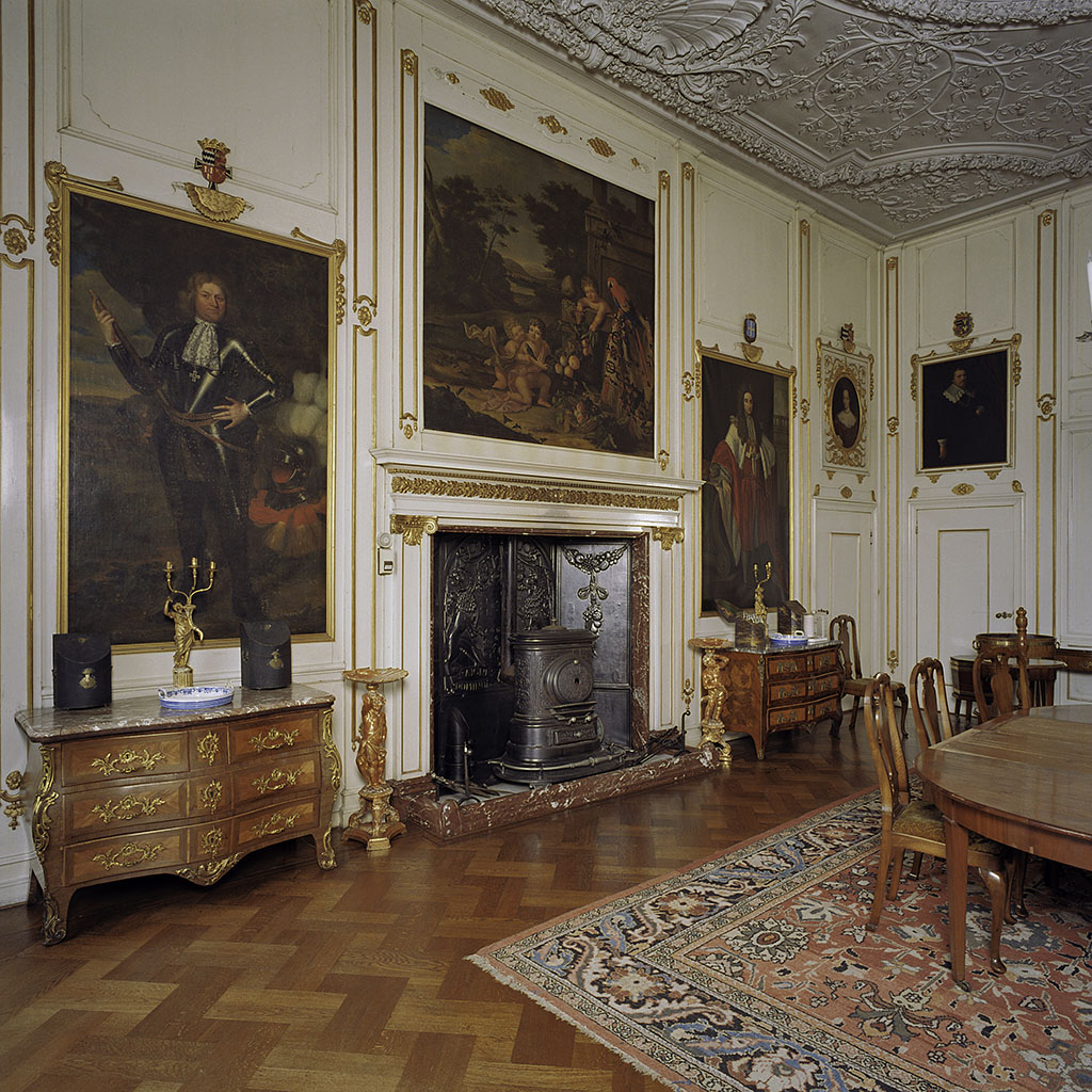 Foto van een eetkamer met rechts een hoek van de eettafel op een vloerkleed. Links een lange wand met beige lambrisering met gouden details. In de lambrisering zijn schilderijen opgenomen. In het midden de schouw met daarin een zwarte gietijzeren kachel, daarboven een schouwstuk met putti, een papagaai en vruchten. Links daarvan een man in een hanrnas, rechts van de schouw een man in een rode mantel. Daarnaast in de hoek een portret van een vrouw en daarnaast in de andere wand het portret van een man. Beide portretten zitten boven deuren. Onder de grote portretten staan comodes en aan weerszijden van de schouw gueridons.