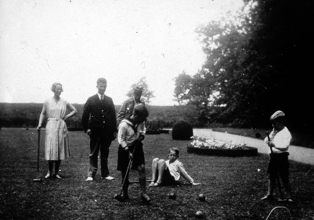 Op een grasveld zijn drie jongens bezig met het spelen van croquet. Een jongen is aan de bal, de ander kijkt geïnteresseerd toe, de derde zit op de grond. Een man en een vrouw kijken toe, op de achtergrond een oudere heer.