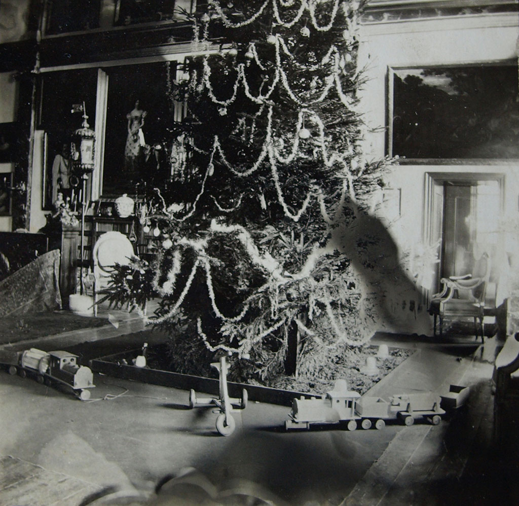 Zwartwit foto van de onderkant van een gigantische kerstboom op de galerij. De boom is versierd met slingers en ballen en eromheen staat speelgoed: een driewieler en twee houten treinen.