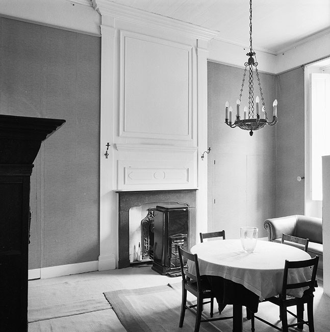 Zwartwitfoto van een vrij kleine kamer met een schouw met daarvoor een vierkante kachel. Rechts een raam. Links een kast, rechts voor het raam een bank en in het midden een ronde tafel met vier stoelen.