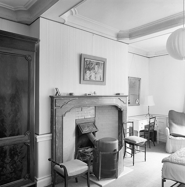 Zwartwitfoto van een kamer met een plafond onderverdeeld in vlakken. Een grote brede schouw met daarvoor een ronde kachel. Links naast de schouw een grote kast, recht een bureautje met daarop een schemerlamp en een spiegel aan de muur. Naast het bureautje een leunstoel. Op de voorgrond zien we nog net een klein stukje van een bed.